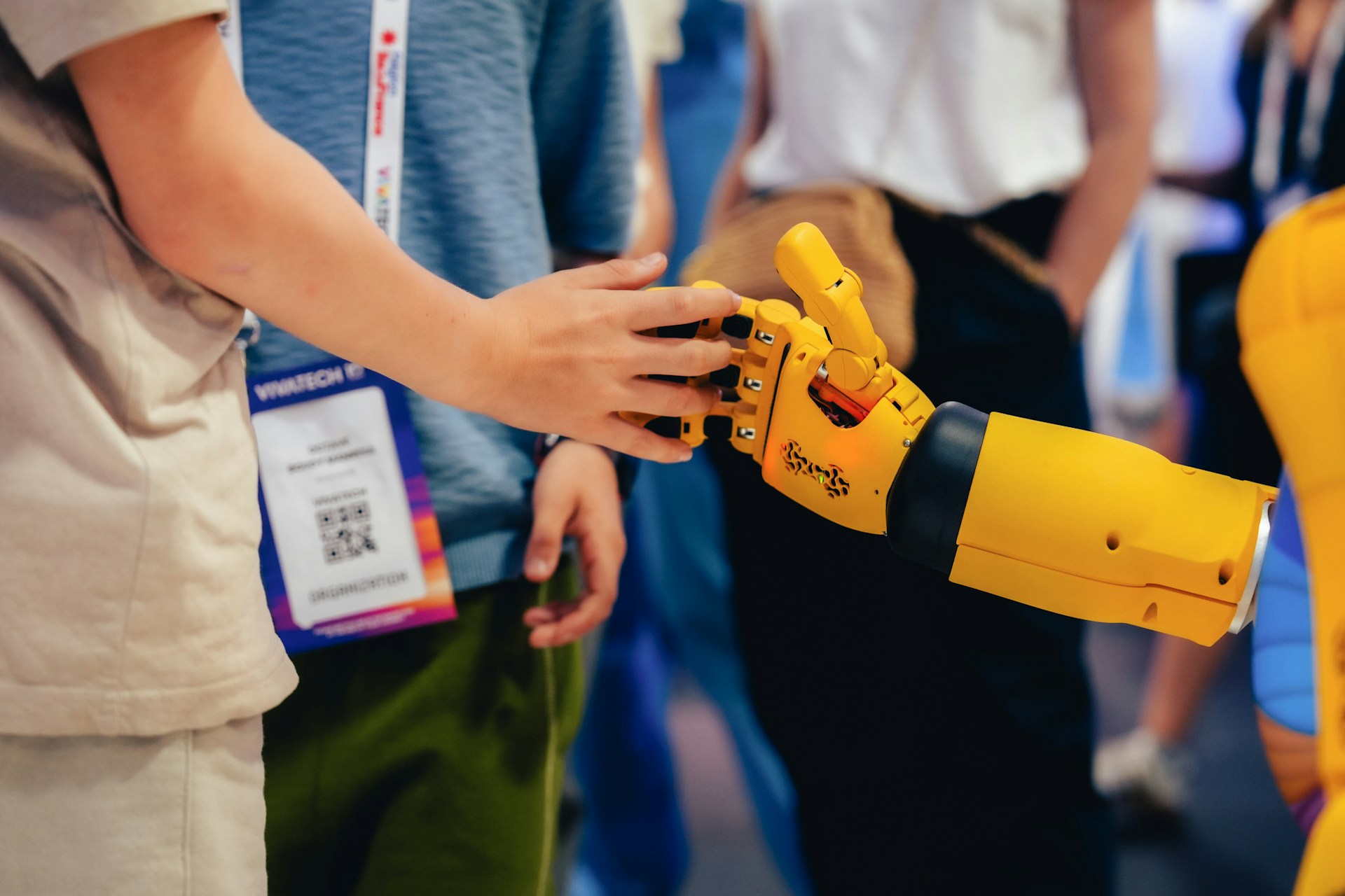 Eine Person berührt die Hand eines gelben Roboters auf einer Technologiemesse, was menschliche Kontrolle über KI darstellt